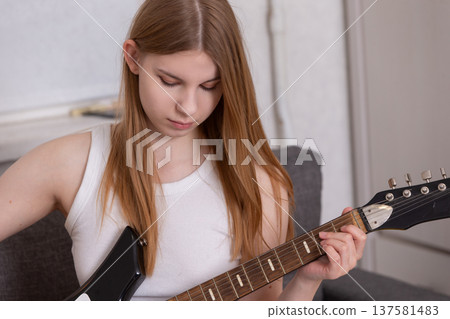 Young woman practicing guitar indoors while seated on a couch in a cozy living room 137581483