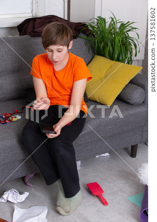 Boy in orange shirt sits on couch using smartphone in a messy living room Boy in orange shirt sits on couch using smartphone in a messy living room 137581602