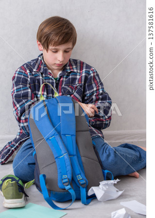 Boy sits on the floor unpacking school supplies from a blue backpack in a light-colored room Boy sits on the floor unpacking school supplies from a blue backpack in a light-colored room 137581616