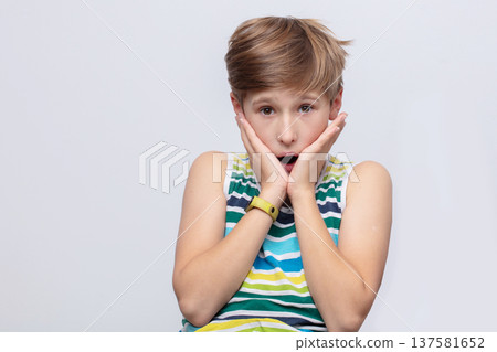 Young boy shows surprise with wide eyes and hands on cheeks, dressed in colorful striped shirt on plain background Young boy shows surprise with wide eyes and hands on cheeks, dressed in colorful striped shirt on plain background 137581652
