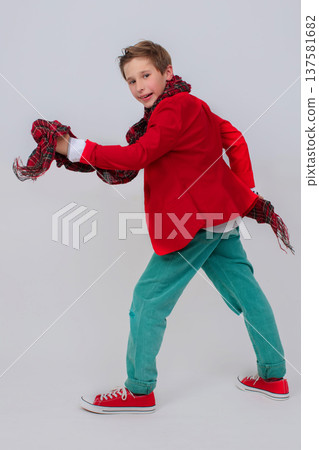 Funny boy in a red jacket, green jeans and sneakers in full growth on a gray background. He takes a big step with his leg up. The concept is only forward. Funny boy in a red jacket, green jeans and sneakers in full growth on a gray background. He takes a big step with his leg up. The concept is only forward. 137581682