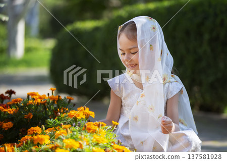 Young girl in a white scarf smiles while tending to orange marigold flowers in a sunny garden in spring Young girl in a white scarf smiles while tending to orange marigold flowers in a sunny garden in spring 137581928
