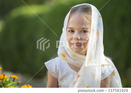 Young girl wearing a scarf smiles in a garden during daylight, surrounded by blooming flowers 137581929