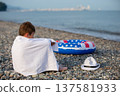A boy wrapped in a towel sits on a pebble beach near a float in the colors of the American flag and a hat on a quiet morning by the sea. 137581933