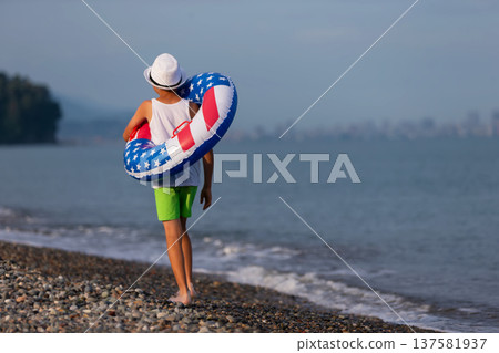 A boy walks along the beach with an inflatable ring in the colors of the American flag on a summer morning near the shore A boy walks along the beach with an inflatable ring in the colors of the American flag on a summer morning near the shore 137581937