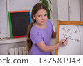 Young girl joyfully solving math problems on a chalkboard in a bright classroom setting 137581943