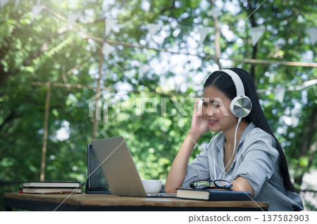 Asian woman wearing headphones listening to music on her laptop. Relaxing time. Lazy worker wasting time at workplace in co-working. Asian woman wearing headphones listening to music on her laptop. Relaxing time. Lazy worker wasting time at workplace in co-working. 137582903