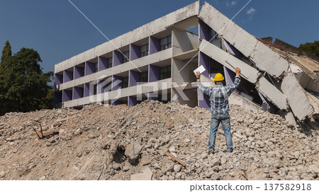 An engineer standing with a tablet and a bluprint raising his hand on pile of collapsed building debris on demolish building. 137582918