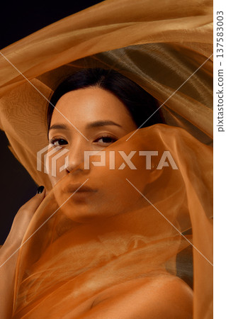 Asian woman cinematic portrait with face partially hidden behind translucent golden silk fabric. 137583003