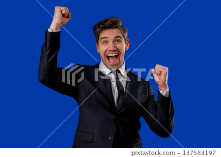 Businessman in suit is very glad and happy, he shows yes gesture of victory with clenched fist, guy achieved result, goals. Surprised excited happy guy on blue background Businessman in suit is very glad and happy, he shows yes gesture of victory with clenched fist, guy achieved result, goals. Surprised excited happy guy on blue background 137583197