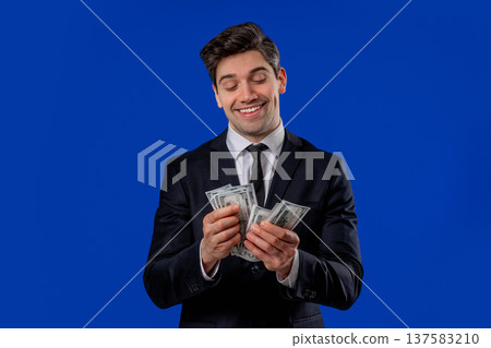 Happy excited businessman counts cash money - USD currency dollars banknotes on blue. Symbol of jackpot, gain, victory, winning the lottery Happy excited businessman counts cash money - USD currency dollars banknotes on blue. Symbol of jackpot, gain, victory, winning the lottery 137583210