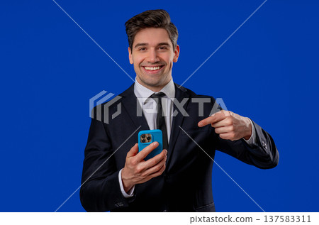 Businessman Pointing at Smartphone Screen on Blue Background Businessman Pointing at Smartphone Screen on Blue Background 137583311