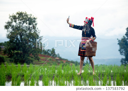 Asian hill tribe woman in traditional ethnic costume taking selfie with smartphone in green rice field. Cultural lifestyle in rural mountain village with terraced rice fields and scenic landscape Asian hill tribe woman in traditional ethnic costume taking selfie with smartphone in green rice field. Cultural lifestyle in rural mountain village with terraced rice fields and scenic landscape 137583792