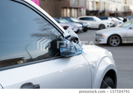 Damaged car side mirror wrapped in a blue plastic bag on a silver vehicle parked in a city street. 137584791