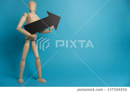 Wooden figure holds black arrow pointing upward against blue backdrop in simple display of direction and movement Wooden figure holds black arrow pointing upward against blue backdrop in simple display of direction and movement 137584850