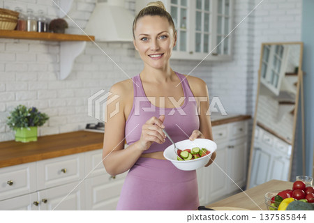 Sporty fit woman smiling in modern kitchen holding bowl with salad, wearing fitness apparel, cooking Sporty fit woman smiling in modern kitchen holding bowl with salad, wearing fitness apparel, cooking 137585844