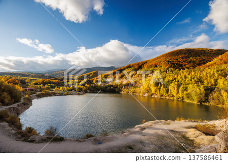 A lake nestled between autumn hills, with golden trees reflecting in the water under a bright blue sky and scattered clouds. Flooded quarry near the village Sutovo in Slovakia, Europe. 137586461