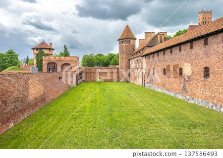 The Malbork Castle, a brick Gothic castle in northern Poland, Europe. Courtyard View of Historic Fortress: Brick walls, lush grass, towers under cloudy skies create an imposing architectural scene. 137586493