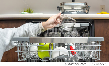 Hand placing glass cup into modern dishwasher filled with clean dishes in a contemporary kitchen setting with wooden cabinetry 137587776