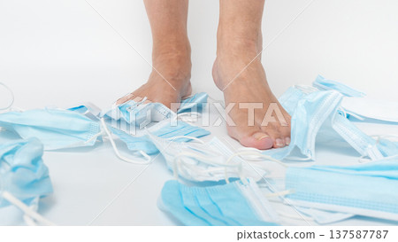 Bare feet standing on discarded blue medical masks scattered on a clean surface, symbolizing the end of a health crisis and environmental impact 137587787