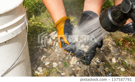 Hands wearing gloves are washing gravel with a hose, showcasing the process of cleaning stones in a natural outdoor setting 137587791