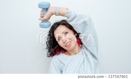 Woman exercising at home with a light blue dumbbell, wearing a gray sweatshirt, against a plain white background with copy space 137587801