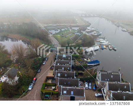 Aerial view of a misty harbor in Muiden, Netherlands. Features a traditional drawbridge, canal with yachts, and residential houses shrouded in thick morning fog. Atmospheric Dutch landscape. Aerial view of a misty harbor in Muiden, Netherlands. Features a traditional drawbridge, canal with yachts, and residential houses shrouded in thick morning fog. Atmospheric Dutch landscape. 137587808
