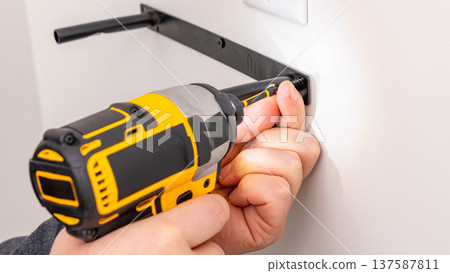 Mans hands operating yellow cordless drill attaching metal bracket to white wall with screw. Home improvement, securing fixture, DIY mounting process, tool in use. 137587811