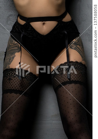 Model poses in black lingerie with straps and thigh-high stockings against a gray wall in an indoor setting 137588058