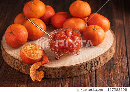 Fresh tangerines on wooden platter accompanied by stemmed glass filled with textured tangerine jam and golden spoon on brown wooden table Fresh tangerines on wooden platter accompanied by stemmed glass filled with textured tangerine jam and golden spoon on brown wooden table 137588306
