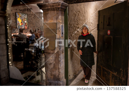Lyon, france. December 28, 2023. Woman in winter jacket and beanie entering a traditional stone-walled restaurant through a heavy wooden door, warm light inviting from inside Lyon, france. December 28, 2023. Woman in winter jacket and beanie entering a traditional stone-walled restaurant through a heavy wooden door, warm light inviting from inside 137588325