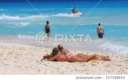 Two women sunbathe on sandy beach while other visitors wade in clear turquoise water Two women sunbathe on sandy beach while other visitors wade in clear turquoise water 137589654