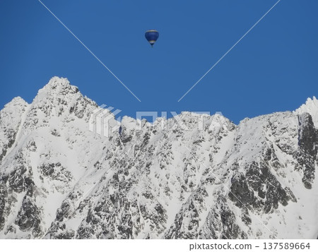 Hot air balloon drifting above rugged snow-covered mountain peaks under a clear blue sky, 137589664