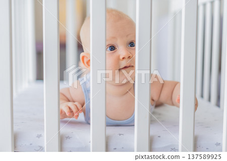 Sad baby looking through the bars of a crib, expressing longing and curiosity. Emotional scene symbolizing the need for freedom and exploration in early childhood. Infant development, autonomy and Sad baby looking through the bars of a crib, expressing longing and curiosity. Emotional scene symbolizing the need for freedom and exploration in early childhood. Infant development, autonomy and 137589925