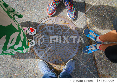 Sakata, Yamagata, Japan - Oct 5 2024, A close-up of a manhole cover with a metal pattern picture in shape of a lighthouse, sea waves, and ship, with legs of a family of four around, Sakata, Japan 137590996