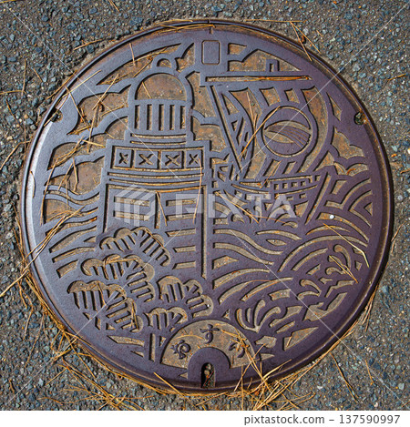 Sakata, Yamagata, Japan - Oct 5 2024, A close-up of a manhole cover with a metal pattern picture in the shape of a lighthouse, sea waves, and ship, on a road in the city of Sakata, Japan  137590997