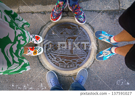 Sakata, Yamagata, Japan - Oct 5 2024,  close-up view of manhole cover with an image of sailing ship sailing down river against of Sankyo Soko Storehouse, legs of family of four around, Sakata, Japan 137591027