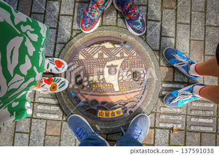 Sakata, Yamagata, Japan - Oct 5 2024, close-up view of a manhole cover with an colored image of ship sailing down river against of Sankyo Soko Storehouse, legs of family of four around, Sakata, Japan 137591038
