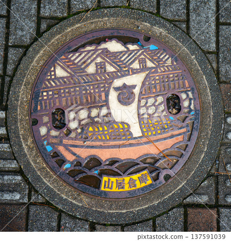 Sakata, Yamagata, Japan - Oct 5 2024, A close-up view of a manhole cover with an colored image of a sailing ship sailing down river against of Sankyo Soko Storehouse, Sakata, Japan Sakata, Yamagata, Japan - Oct 5 2024, A close-up view of a manhole cover with an colored image of a sailing ship sailing down river against of Sankyo Soko Storehouse, Sakata, Japan 137591039