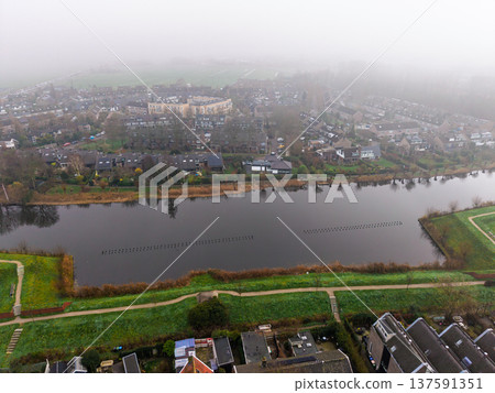 Moody aerial view of a Dutch town by a canal in thick fog. Green embankments and historic residential houses shrouded in morning mist. Atmospheric European landscape in autumn or winter. Moody aerial view of a Dutch town by a canal in thick fog. Green embankments and historic residential houses shrouded in morning mist. Atmospheric European landscape in autumn or winter. 137591351
