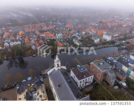High angle aerial view of a historic Dutch town featuring a white church and red roofed houses along a canal. Thick morning fog creates a moody, atmospheric European landscape. 137591353