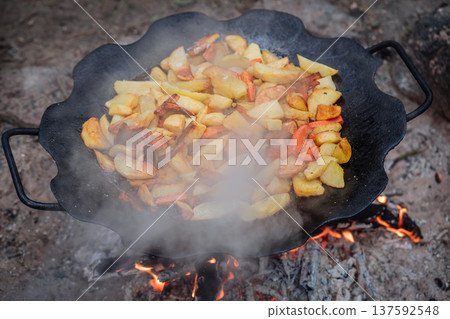 Fried potatoes with carrots cooking in a cast iron pan over an open campfire outdoors 137592548