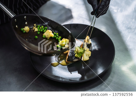 Professional chef plating grilled fish with broccoli and green peas using tweezers in restaurant kitchen Professional chef plating grilled fish with broccoli and green peas using tweezers in restaurant kitchen 137592556