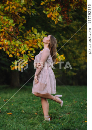 Beautiful young woman in a pink dress posing in autumn park with yellow trees on background Beautiful young woman in a pink dress posing in autumn park with yellow trees on background 137592586