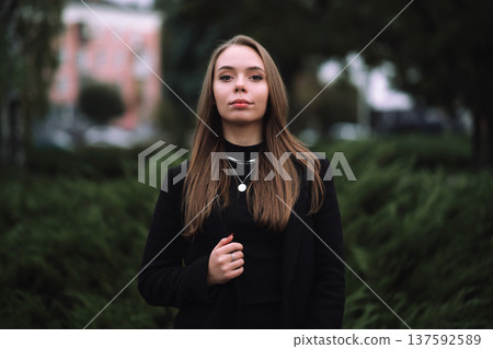 Young stylish woman in black coat and silver jewelry posing in city park outdoors 137592589