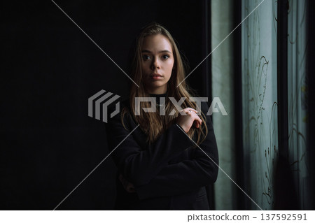 Low key portrait of a mysterious young woman in a black jacket posing near a window in the dark Low key portrait of a mysterious young woman in a black jacket posing near a window in the dark 137592591