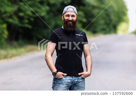Smiling brutal man with a long beard and mustache wearing a cap and black polo shirt outdoors Smiling brutal man with a long beard and mustache wearing a cap and black polo shirt outdoors 137592600
