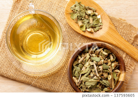 A glass cup of lemon balm tea, surrounded by dried leaves in a bowl, and a wooden spoon placed on burlap fabric 137593605