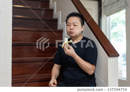 A man sits on the stairs at home, smiling while eating a banana as a healthy snack and source of natural energy after exercise. A man sits on the stairs at home, smiling while eating a banana as a healthy snack and source of natural energy after exercise. 137594759