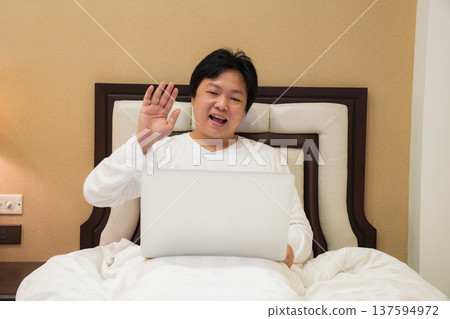 A man waves while talking online from his bedroom. The relaxed atmosphere shows comfort, connection, and modern communication at home. A man waves while talking online from his bedroom. The relaxed atmosphere shows comfort, connection, and modern communication at home. 137594972